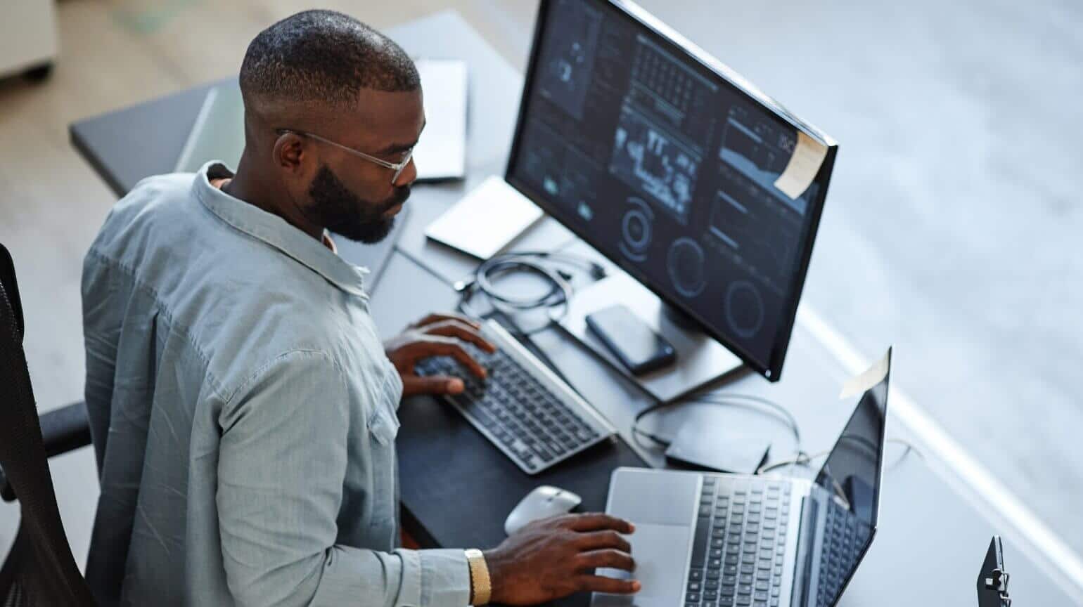 Man working at a desk on a laptop with a monitor displaying data